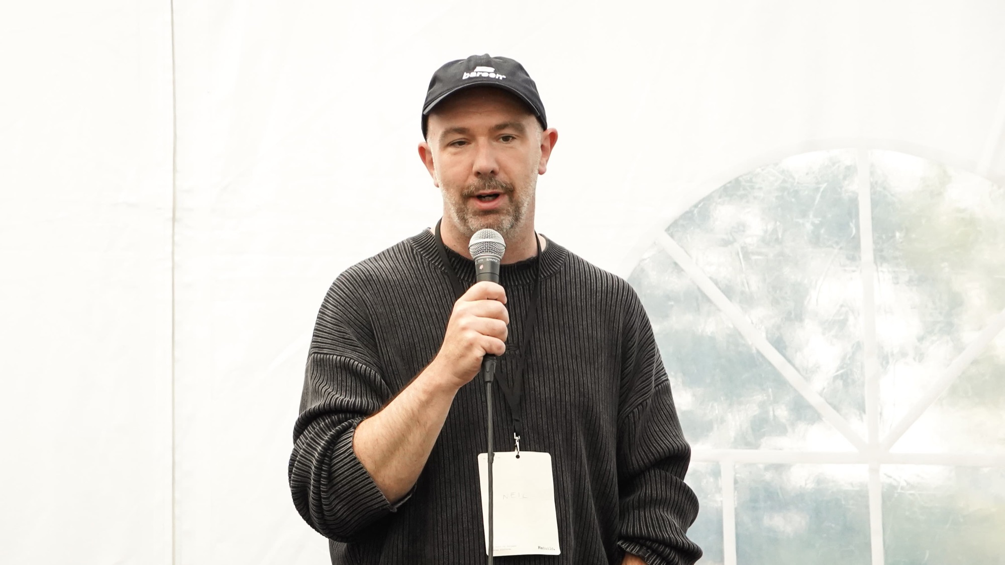 A speaker in a black cap and lanyard talks into a microphone inside the Rebuild2 tent.