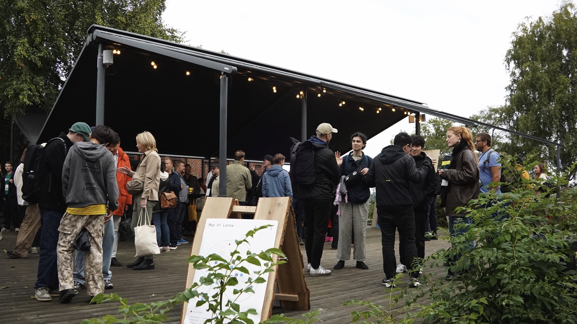 Attendees gather and talk on a wooden deck under a covered pavilion strung with lights, beside a “Map of Lonna” sign.
