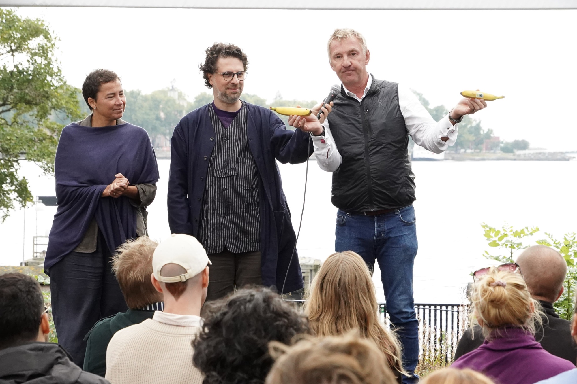 Three speakers stand before a seated audience by the water, two of them holding up bananas.