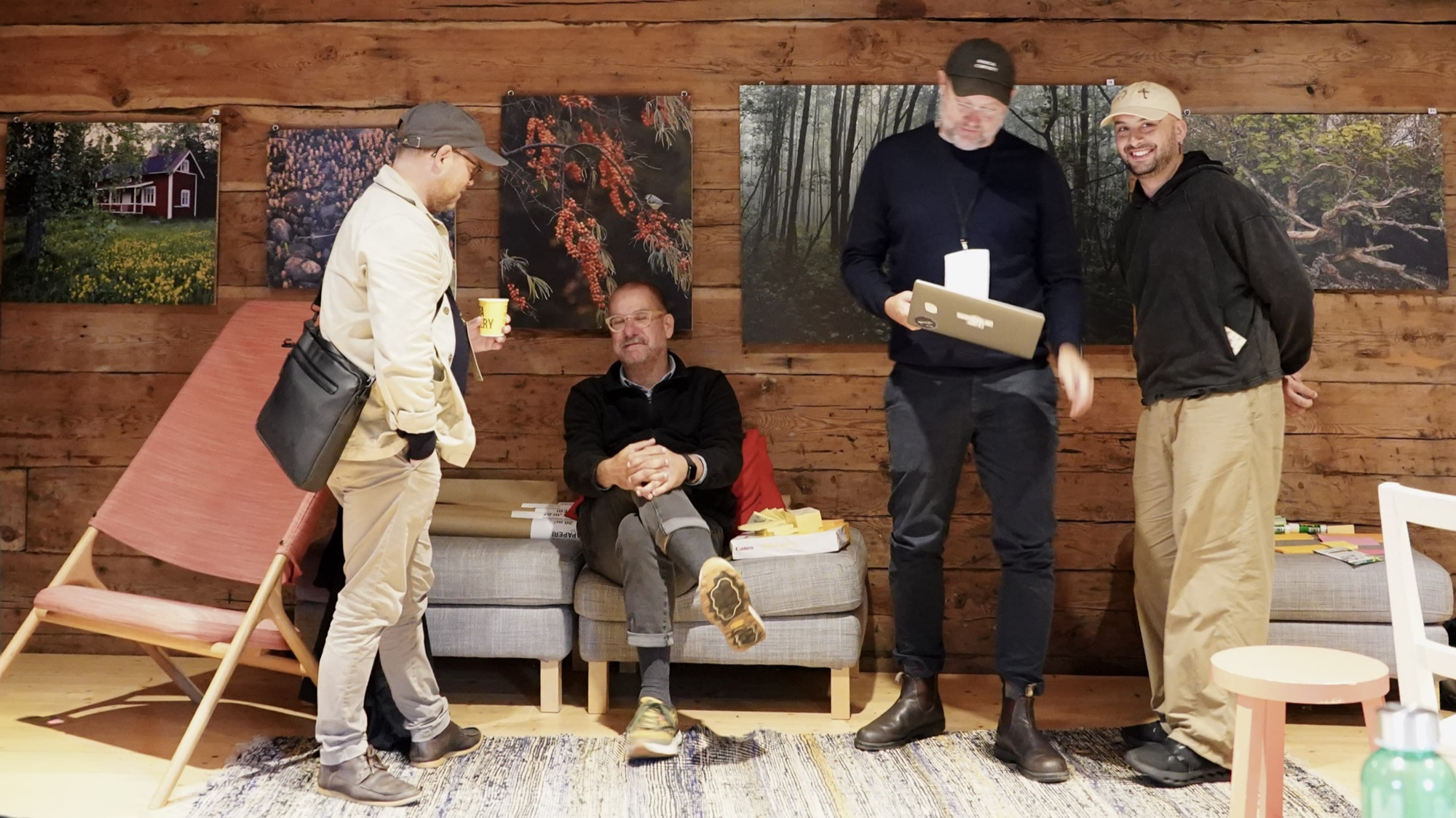 Four people talk in a timber-walled room hung with nature photographs, one holding a laptop.