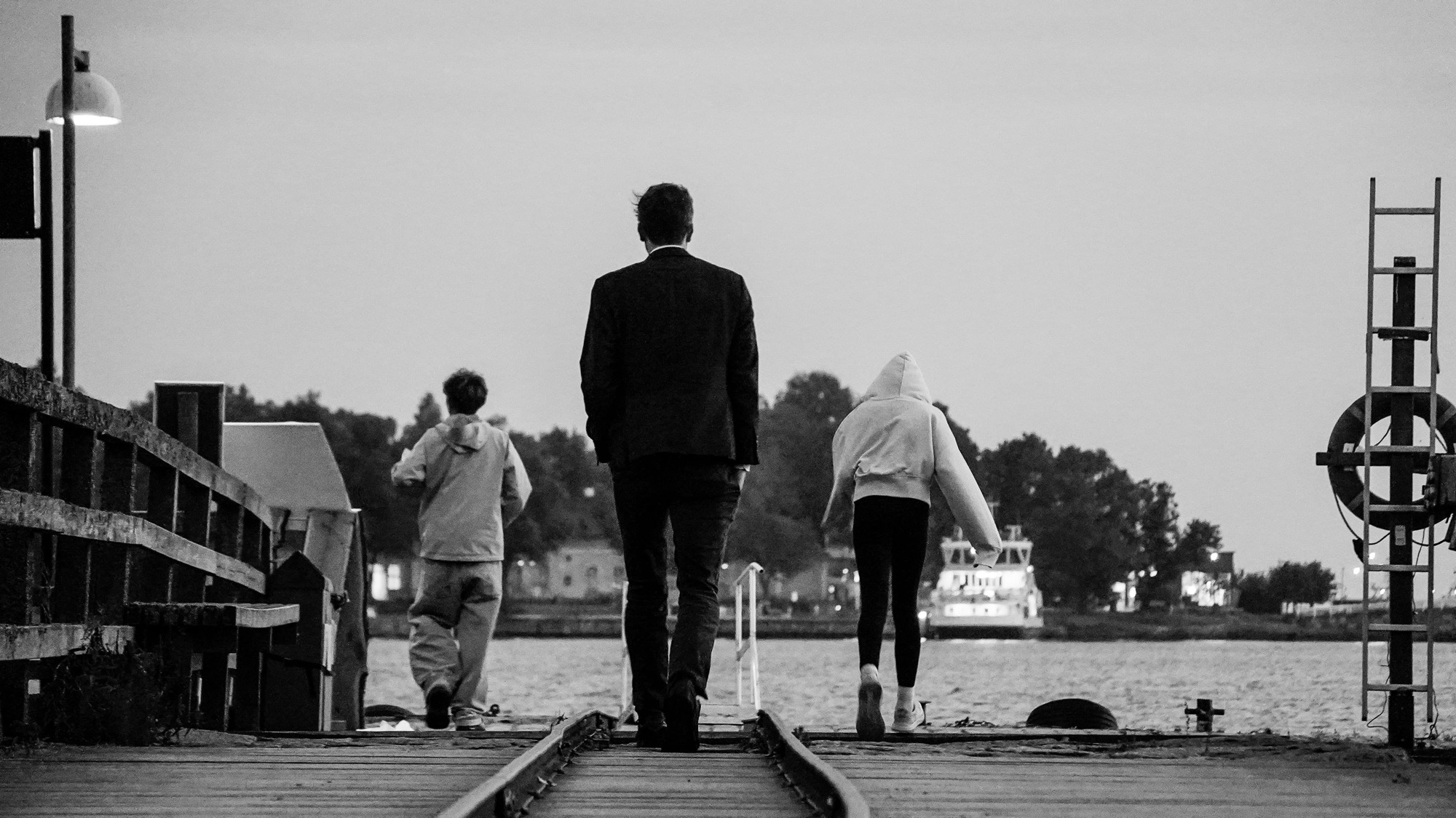 Three people walk away from the camera along a wooden pier towards the water at dusk, in black and white.